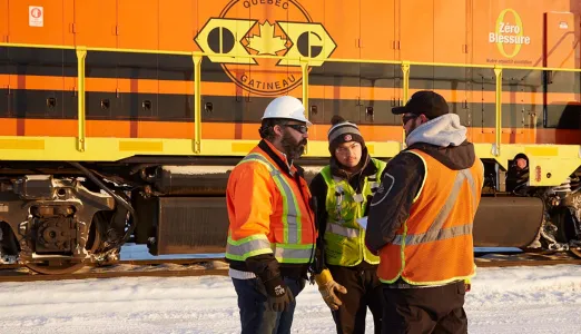 Three G&W employees wearing safety equipment and high-visibility clothing huddled talking to each other in front of a train in a snowy landscape