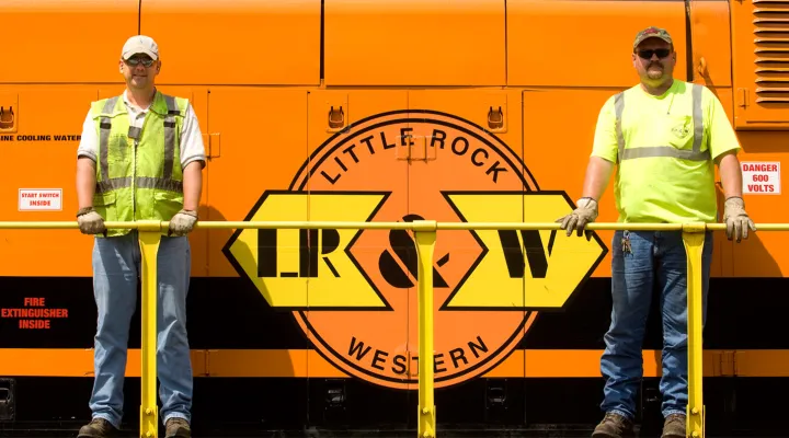 Two G&W employees wearing high-visibility clothing, caps, and sunglasses standing behind the railing on the site of a train next to the Little Rock & Western Railway logo